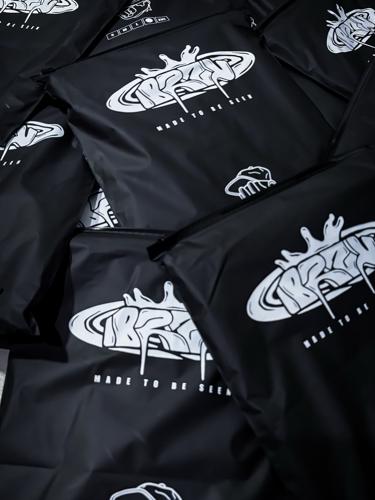 Black zippered pouches or bags with a white graffiti-style logo and the text 'MADE TO BE SEEN' printed on them, shown in a close-up pile.
